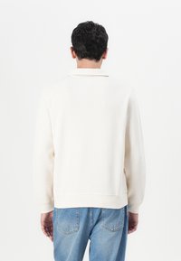 Personne aux cheveux courts et foncés portant une chemise écrue à manches longues et un jean bleu, debout face à un fond blanc uni.