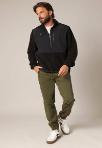 Fleece jack met zwarte en grijze panelen, ritssluiting en een zak aan de voorkant; gecombineerd met groene broek en witte sneakers met zwarte accenten.