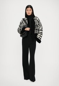 Cardigan surdimensionné à motifs noir et blanc avec une finition texturée, porté sur un col roulé noir et un pantalon évasé. Sac à main noir inclus.