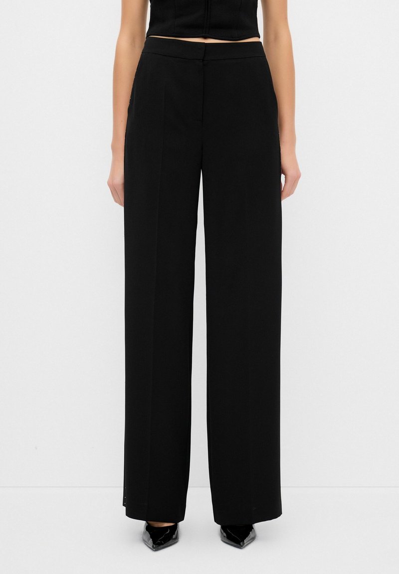 Pantalons larges noirs en tissu lisse, avec une taille ajustée et des poches latérales discrètes. Le design présente des lignes épurées et une silhouette élégante.