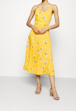 Robe midi jaune avec imprimé floral, bretelles fines, taille cintrée, jupe fluide, tissu lisse, associée à des sandales à talons blocs beiges.