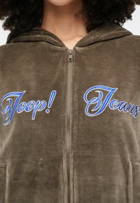 Close-up de uma pessoa vestindo um hoodie verde-oliva com fecho de correr e texto bordado em azul "Joop!" à esquerda e "Jeans" à direita.