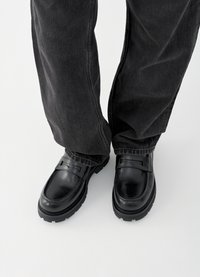 Juodi odiniai loaferiai su lygiu paviršiumi, puošti dekoratyviniu dirželiu per viršų ir tvirtu padu, derinami su tamsiomis kelnėmis.