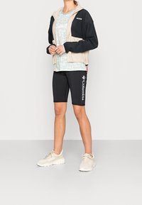 Béžová a černá bunda na zip s kapucí, světle modrá vzorovaná košile, černé šortky s bílým logem a krémové tenisky.