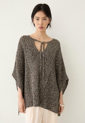 Ung kvinde iført en løs, sort-og-hvid meleret striktrøje med vide ærmer og bindebånd ved halsen, kombineret med lyse bukser.