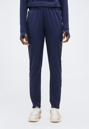 Osoba oblečená v tmavomodrých športových nohaviciach a zodpovedajúcom dlhom rukáve, stojaca s rukami voľne pozdĺž tela, viditeľné biele tenisky.