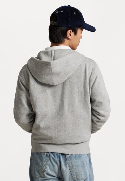 Homem de costas vestindo um moletom cinza, jeans azul e um boné de veludo cotelê azul escuro, com um fundo branco.