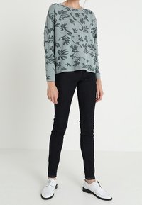 Helles türkisfarbenes Sweatshirt mit botanischem Print, kombiniert mit schwarzen Skinny-Jeans und weißen Schuhen. Das Sweatshirt hat lange Ärmel und einen runden Ausschnitt.