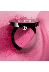 Rund svart kompakt som inneholder lys rosa rouge med en jevn tekstur og preget logo på overflaten, omgitt av rosa pudder.