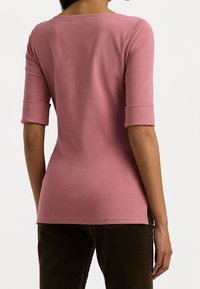 Nainen, joka on pukeutunut tiukkaan pölyruusunväriseen puoleen hihansuun yläosaan, jossa on käänteet, ja tummanoliivinvärisiin housuihin, kuvattuna takaa valkoista taustaa vasten.