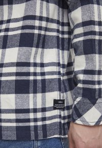 Camicia a quadri blu navy e bianca in cotone, con polsini bottone e un'etichetta nera all'orlo. Design a quadri ben visibile e dettagli sulla texture.