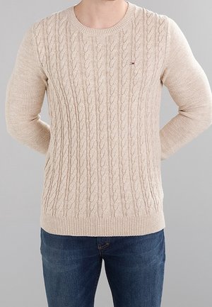Mężczyzna noszący beżowy sweter z grubym splotem i niebieskie dżinsy, stojący z rękami za plecami na jednolitym szarym tle.