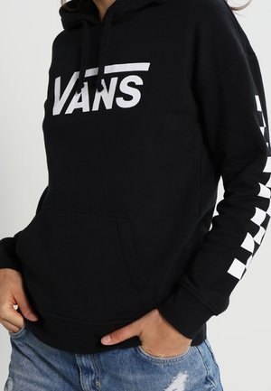 Sort Vans hættetrøje med hvidt logo på brystet og hvidt ternmønster på højre ærme, båret med blå revnede jeans.