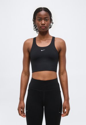 Mladá žena v čiernom tielku bez rukávov a čiernych legínach s vysokým pásom, stojaca na jednoduchom bielom pozadí.