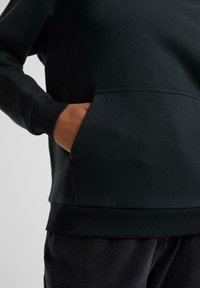 Persoană purtând un hanorac negru, cu mâna stângă în buzunarul din față, arătând manșetele și tivul striate, pe un fundal simplu și deschis la culoare.