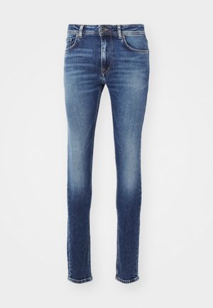 Blugi din denim albastru, cu o croială slim, având talie medie, buzunare cusute și o ușoară decolorare. Tivurile sunt ușor deteriorate.