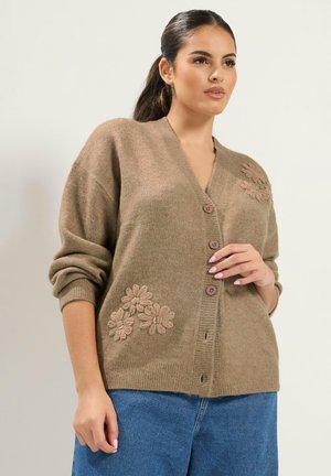 Femme portant un cardigan marron boutonné avec broderies florales et un jean bleu, debout devant un fond clair uni.