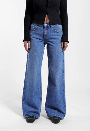 Person wearing high-waisted wide-leg blue jeans, black cropped buttoned sweater, and black shoes with white stitching, standing on white background.