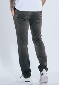 Mann trägt dunkelgraue Slim-Fit-Hosen und weiße Sneakers mit schwarzen Akzenten, steht mit einer Hand in der Tasche und hat einen tätowierten Unterarm sichtbar.