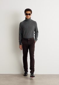 Gray turtleneck sweater, brown slim-fit pants, and black shoes. Model wears dark sunglasses. Plain background, focus on outfit.