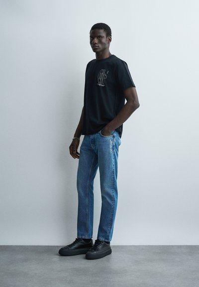 Άνδρας στέκεται με το ένα χέρι στην τσέπη του τζιν, φορώντας μαύρο μπλουζάκι με στάμπα, μπλε τζιν με λογότυπο στο μηρό και μαύρα αθλητικά παπούτσια μπροστά σε λευκό τοίχο.