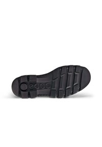 Semelle de chaussure en caoutchouc noir avec une surface texturée, présentant des nervures décoratives et un détail de logo circulaire près du talon.