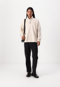 Bežový svetrový pulover na zips, vo voľnom strihu, skombinovaný s čiernymi cargo nohavicami s bočnými vreckami. Čierne topánky dokončujú outfitt.
