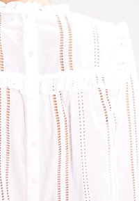 Vit blus med horisontella utskärningar, volangkrage och knappstängning. Lättviktsmaterial med texturerade detaljer.