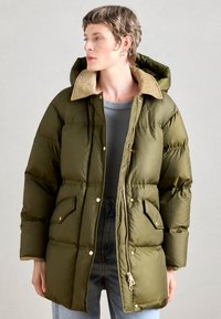 Parka căptușit în verde măsliniu cu o căptușeală din blană artificială bej, având fermoar frontal, capse și buzunare laterale. Țesătură netedă, rezistentă la apă.
