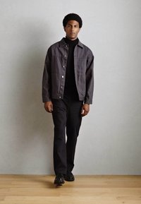 Chaqueta gris oscuro sobre un cuello de tortuga negro, combinada con pantalones negros y zapatos negros, con un fondo claro y texturizado.