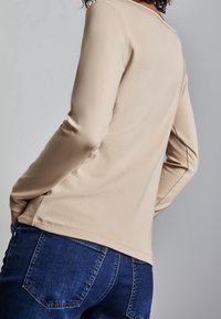 Persona che indossa un top a maniche lunghe beige e jeans di denim blu, con le mani nelle tasche posteriori, leggermente girata lontano dalla fotocamera su uno sfondo grigio.