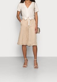 Mujer vistiendo una blusa blanca de manga corta atada, pantalones cortos anchos de longitud a la rodilla en color beige, tacones nude con tiras al tobillo y un pequeño bolso cruzado con estampado de serpiente.