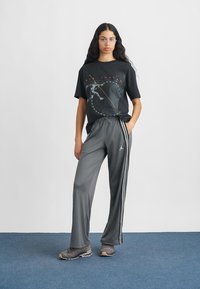 Γκρι φόρμα με ρίγες στο πλάι, συνδυασμένη με μαύρο γραφικό t-shirt με τυπωμένο σχέδιο. Το μοντέλο φορά γκρι αθλητικά παπούτσια.