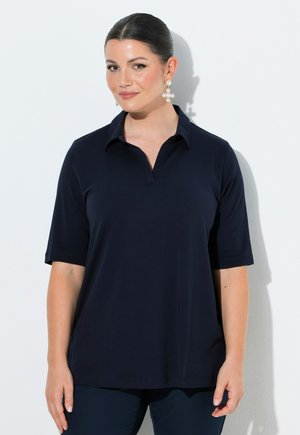 Kvinde med mørkt hår i en lav knold iført en navy kortærmet skjorte med krave og store perlekors-øreringe, stående mod en hvid baggrund.