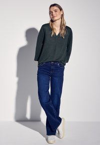 Tummanvihreä V-kauluksinen paita pitkähihaisilla hihoilla, yhdistettynä sinisiin suoriin farkkuihin ja valkoisiin tennareihin, vaaleaa taustaa vasten.