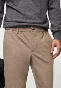 Pantalon beige à texture lisse, doté de poches latérales, d'une fermeture à bouton à l'avant et de passants de ceinture. Porté avec un pull gris foncé.