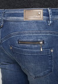 Jeans de mezclilla azul oscuro con un bolsillo con cremallera y una etiqueta de cuero en la cintura. La tela presenta una textura ligeramente deslavada.