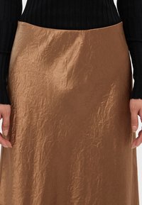 Brun metallisk midi skjørt med rynkete tekstur, høy livlinje, sammen med en svart ribbestrikket langermet topp, hender synlige ved livet.