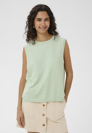 Femme souriante aux cheveux bouclés et foncés, portant un haut sans manches vert clair et une jupe beige boutonnée, debout devant un fond clair uni.