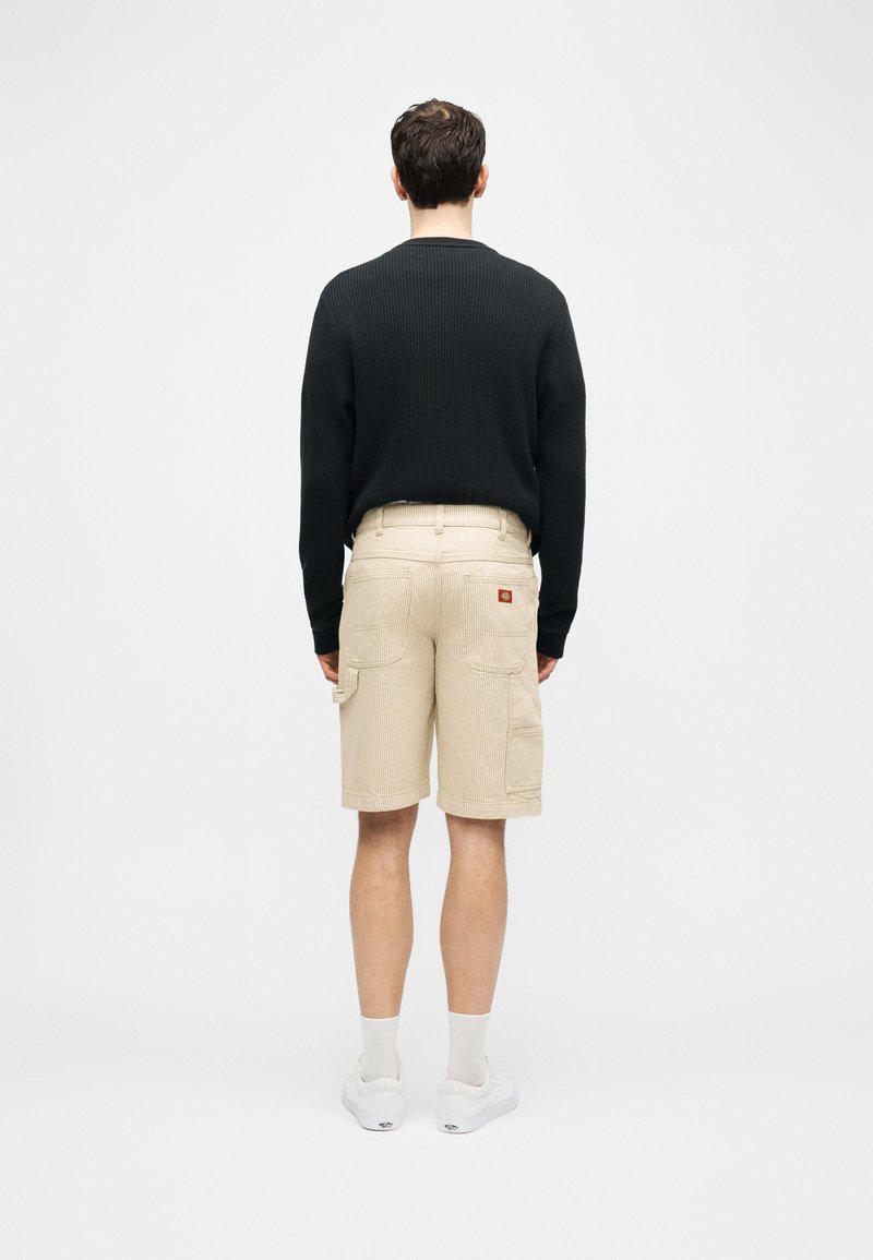 Man die met zijn rug naar de camera staat, gekleed in een zwart shirt met lange mouwen, beige cargoshorts, witte sokken en witte sneakers, tegen een effen grijze achtergrond.