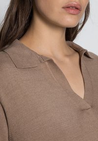 Pull en maille marron avec un col en V, présentant une texture douce et un matériau léger, avec des coutures détaillées au niveau du col.