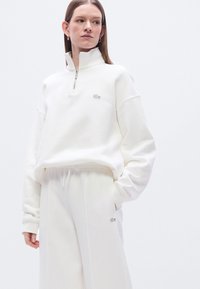 Λευκό φούτερ με φερμουάρ και ψηλό γιακά, ριμπ μανσέτες και μικρό κεντημένο λογότυπο. Συνδυάζεται με αντίστοιχο λευκό παντελόνι φόρμας.