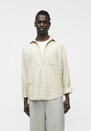 Młody mężczyzna w jasnobeżowej koszuli w kratę noszonej na białym t-shircie, w luźnych, jasnoszarych spodniach, pozujący neutralnie.