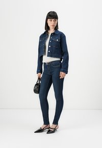 Woman with short dark hair wearing cropped denim jacket, white top, dark skinny jeans, black pointed shoes, and holding a small black handbag.