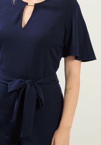 Tuta blu navy con scollatura a chiave, maniche svolazzanti e cintura annodata in vita. Realizzata in tessuto liscio con una leggera lucentezza.