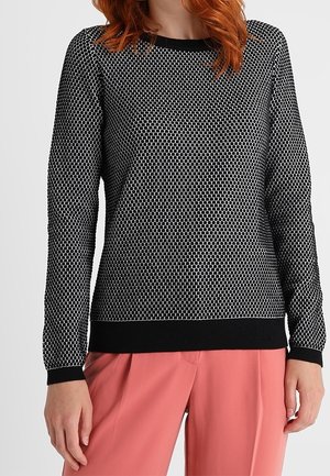 Femme portant un pull à manches longues à motifs noir et blanc avec des poignets noirs et un pantalon large couleur corail rose.