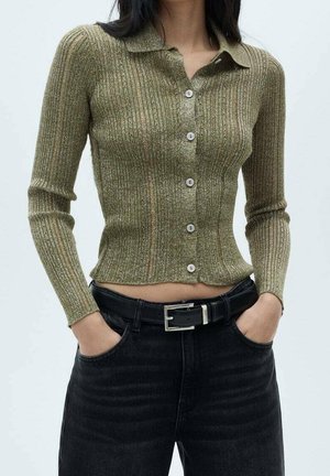 Vrouw draagt een nauwsluitende, geribbelde, olijfgroene cardigan met knopen en een hoge taille zwarte spijkerbroek, met handen in de zakken, tegen een effen achtergrond.