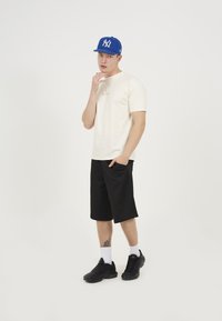 Cream t-shirt, black shorts, blue cap with logo, black sneakers, white socks. Standing casual pose, one hand on chin, neutral background.