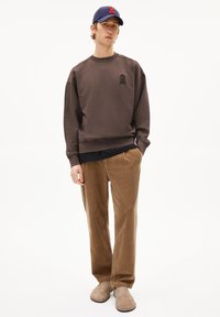 Sweatshirt marron à col rond avec des détails brodés, associé à un pantalon en velours côtelé beige et des chaussures à enfiler beiges. Casquette navy avec des accents rouges.