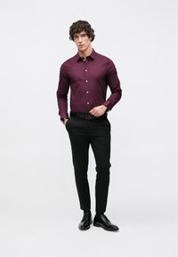 Camisa de manga larga color burdeos con diseño ajustado, combinada con pantalones negros y zapatos negros de cordones, posando frente a un fondo neutro.